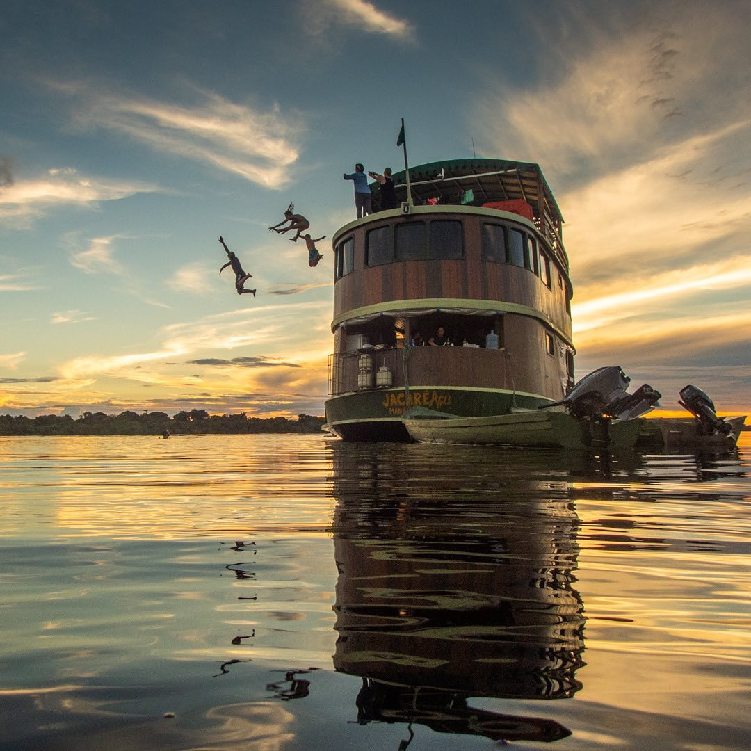 Tourists jumping into the river from the top deck of the Jacaré-Açu wooden boat at sunset during an authentic Amazon river cruise experience. Tourists jumping into the river from the top deck of the Jacaré-Açu wooden boat at sunset during an authentic Amazon river cruise experience.