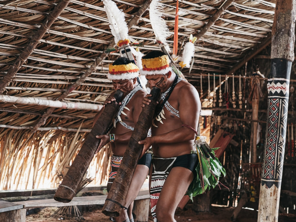 Two men from the Tuyuka indigenous community wear feathered headdresses and traditional garments while playing large wooden flutes.
